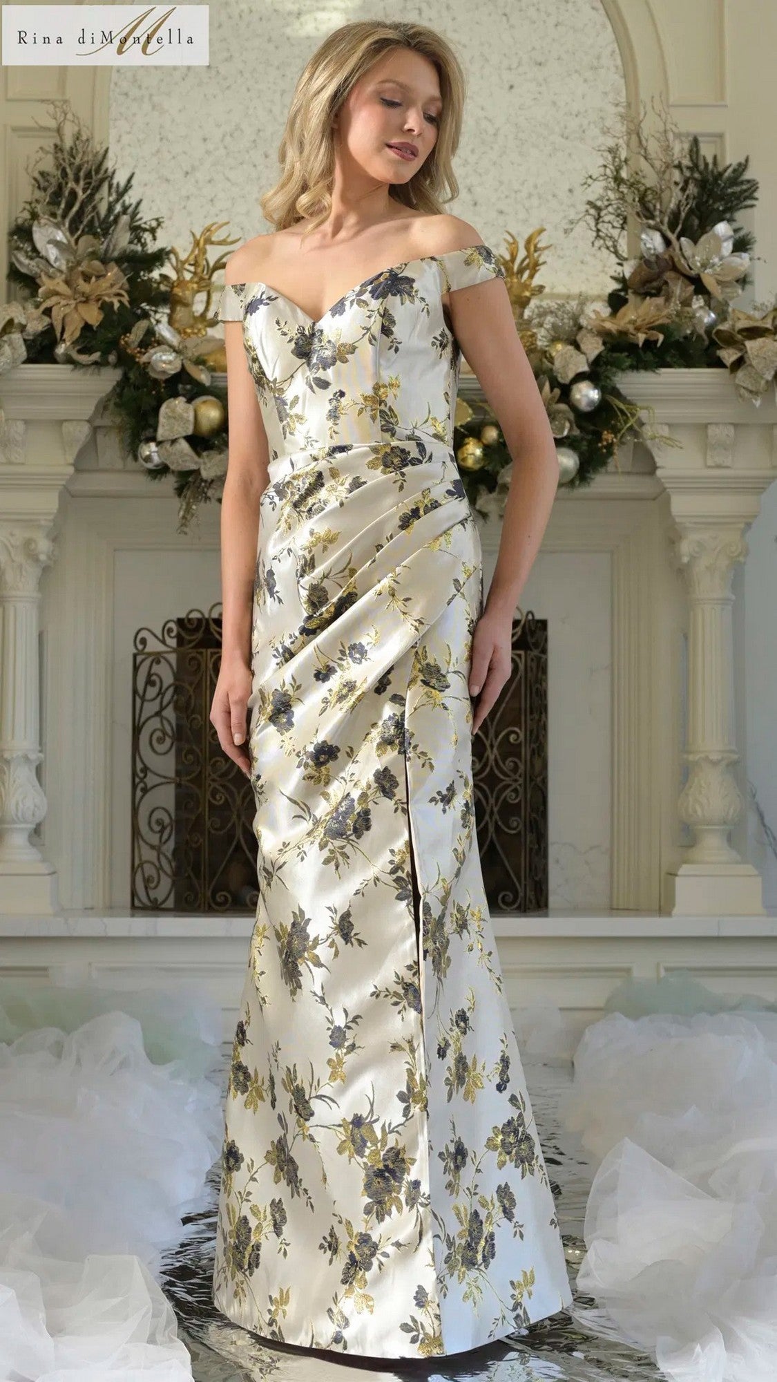 A woman wears the Rina Di Montella RD3350 long formal dress in Navy Multi with off-the-shoulder sleeves, floral print, and a front slit, standing indoors before a decorated white fireplace.